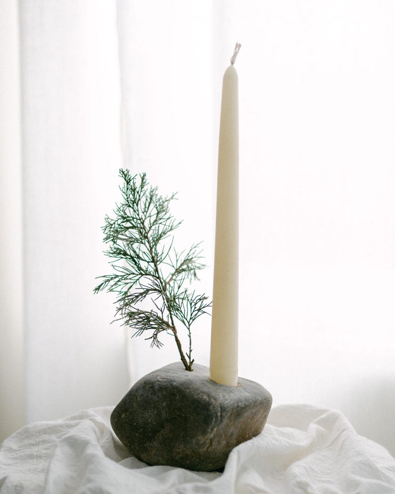 May include: A natural stone candle holder with a cream-colored taper candle and a small evergreen sprig. The candle holder is a dark gray color and sits on a white cloth. The candle is approximately 10 inches tall.