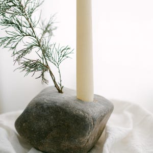 May include: A gray stone candle holder with a cream-colored candle and a small green sprig. The candle holder is a natural, rustic design, perfect for home decor. The candle is a standard size.