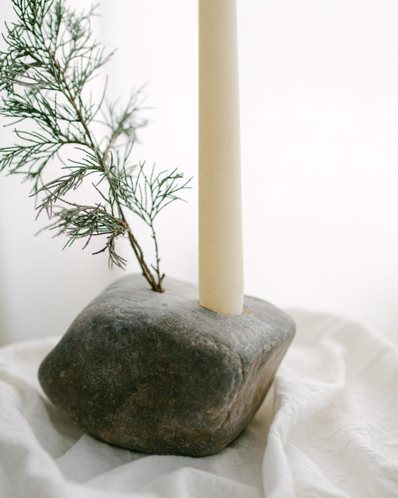 May include: A gray stone candle holder with a cream-colored candle and a small green sprig. The candle holder is a natural, rustic design, perfect for home decor. The candle is a standard size.