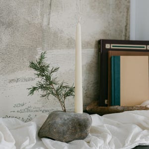 May include: A natural stone candle holder with a tall, cream-colored taper candle and a sprig of greenery. The candle holder sits on a white linen cloth, with books in the background. The candle is approximately 10 inches tall.