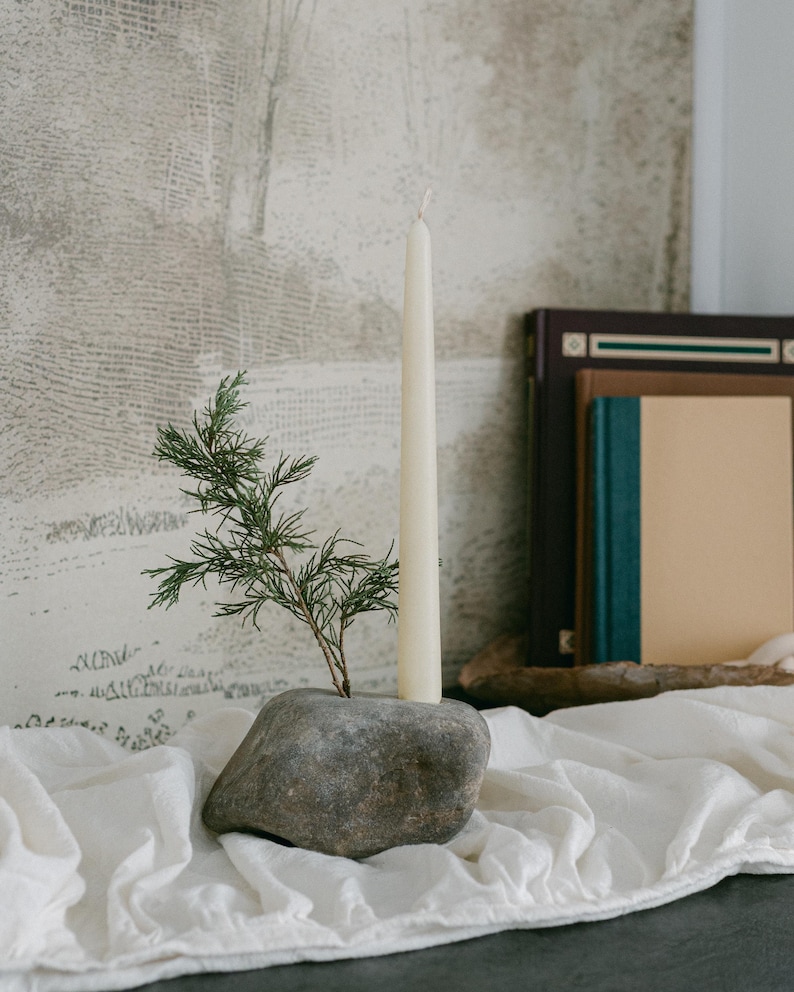 May include: A natural stone candle holder with a tall, cream-colored taper candle and a sprig of greenery. The candle holder sits on a white linen cloth, with books in the background. The candle is approximately 10 inches tall.