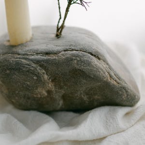 May include: A gray, natural stone candle holder with a cream-colored candle and a small sprig of greenery. The stone has a rough texture and is set on a white, textured fabric. The candle holder is a unique, rustic home decor item.
