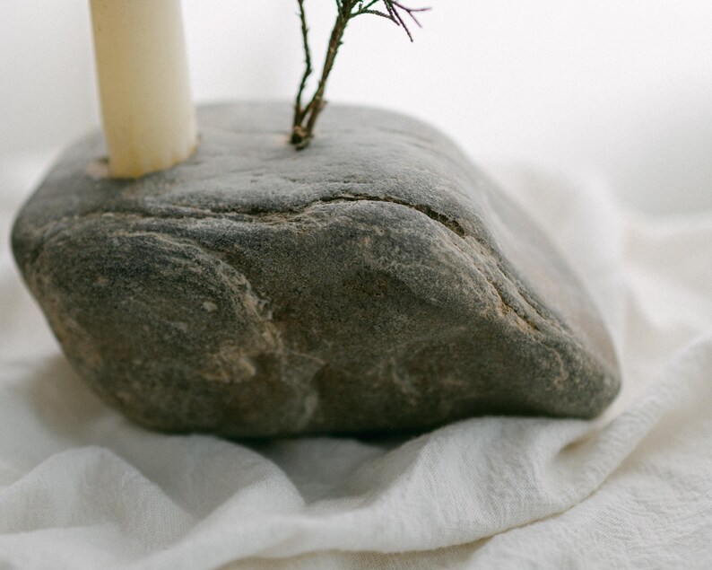 May include: A gray, natural stone candle holder with a cream-colored candle and a small sprig of greenery. The stone has a rough texture and is set on a white, textured fabric. The candle holder is a unique, rustic home decor item.