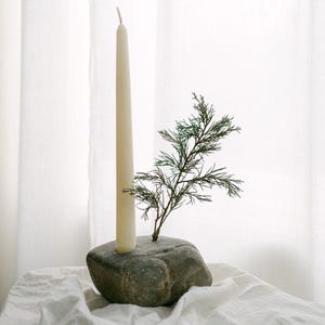 May include: A cream-colored taper candle is set in a gray stone candle holder. A small green sprig of foliage is placed next to the candle. The candle holder sits on a white cloth.