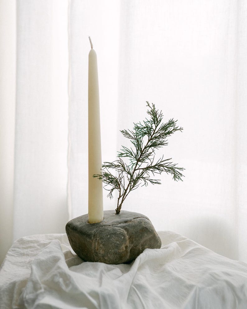 May include: A cream-colored taper candle is set in a gray stone candle holder. A small green sprig of foliage is placed next to the candle. The candle holder sits on a white cloth.