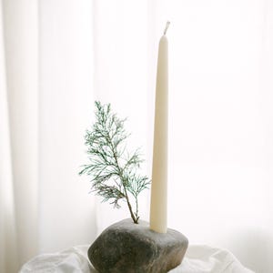 May include: A natural stone candle holder with a cream-colored taper candle and a small evergreen sprig. The candle holder is sitting on a white cloth, creating a minimalist and earthy aesthetic.