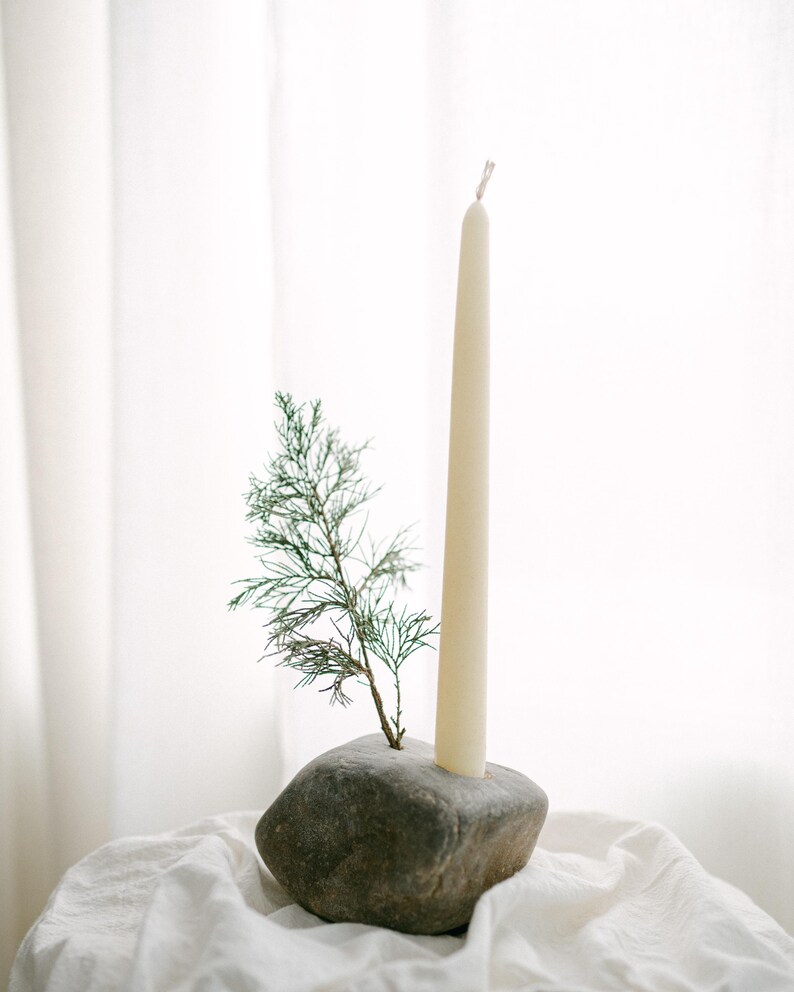 May include: A natural stone candle holder with a cream-colored taper candle and a small evergreen sprig. The candle holder is sitting on a white cloth, creating a minimalist and earthy aesthetic.