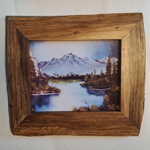 May include: A landscape painting of snow-capped mountains reflecting in a calm lake, surrounded by evergreen trees and golden foliage. The artwork is encased in a rectangular wooden frame featuring a natural, rough-hewn live edge finish.