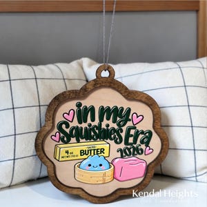 May include: A scalloped wooden ornament featuring the text "in my Squishies Era 2026" in dark green script. The design includes a yellow 113 g salted butter stick, a blue dumpling in a bamboo steamer, a pink block, and small pink hearts.