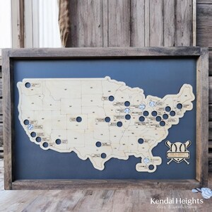 May include: A wooden map of the United States with baseball stadiums marked with blue baseballs. The map is framed in a dark wood frame and has a blue background. The text "Todd & Amber's" is visible in the lower right corner of the map.
