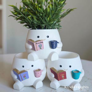 May include: Three white ceramic planters shaped like small figures with arms and legs. Each figure holds a miniature book and a mug. The top figure's book is labelled Grandma with a flower. The planters contain green leafy artificial plants.