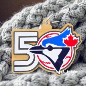 Peut inclure: Ornement en bois avec le chiffre "50" en blanc et un logo de baseball avec un geai bleu et une feuille d'érable rouge. L'ornement a un bord doré et une ficelle argentée pour l'accrocher.