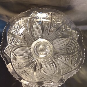 Puede incluir: Un cuenco de cristal transparente con pedestal, borde festoneado y dise&ntilde;o de cristal tallado intrincado. El cuenco tiene un pedestal central con una base circular.