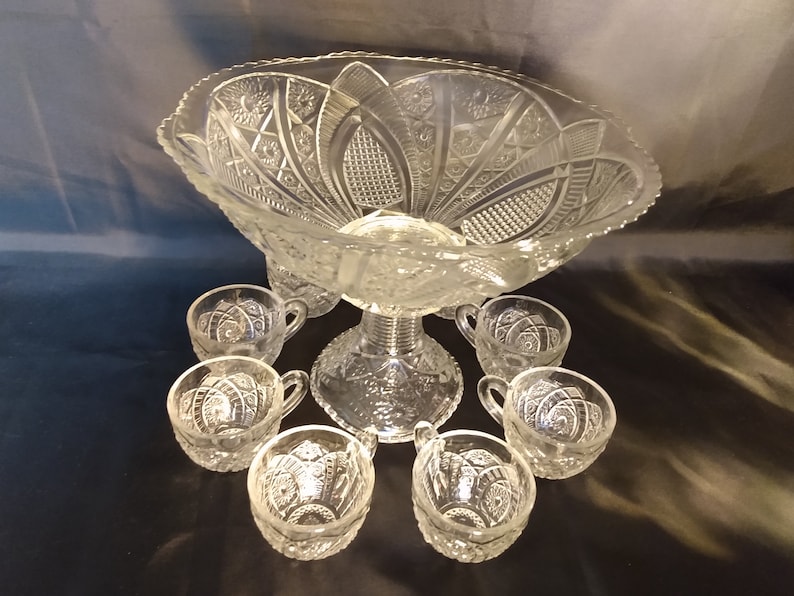Puede incluir: Un juego de ponchera de cristal vintage con un cuenco de cristal transparente y ocho tazas a juego. El cuenco tiene un dise&ntilde;o decorativo de cristal tallado y una base de pedestal. Las tazas tambi&eacute;n son de cristal tallado y tienen un asa.