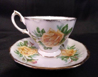 May include: A white teacup and saucer with a gold trim. The teacup and saucer are decorated with yellow roses and green leaves.