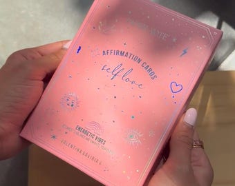 Puede incluir: Caja rosa de cartas de afirmación con el texto "AFFIRMATION CARDS self love" y "ENERGETIC VIBES" en la parte delantera. La caja está decorada con estrellas, corazones y otros símbolos.