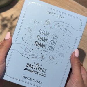 May include: A light blue box with silver lettering that reads "PAPER NOTE" and "THANK YOU" three times. The box contains 30 gratitude affirmation cards by Valentina Gaviría U.