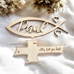 May include: Wooden cutout of a fish with the name "Paul" inside and two hearts. Below is a wooden cross with the text "Alles Liebe zur Taufe".
