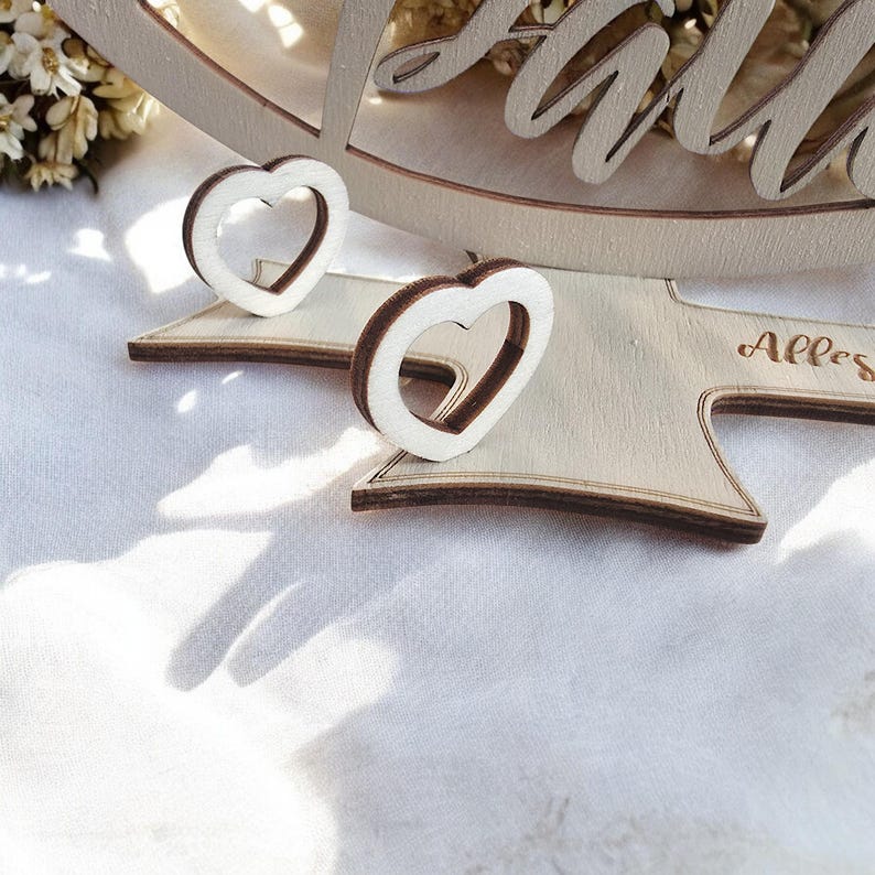 May include: Two wooden heart-shaped ornaments with a cutout design. The ornaments are on a wooden base with the word "Alles" engraved on it.