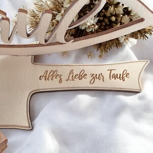 May include: A wooden sign with the words "Alles Liebe zur Taufe" engraved on it. The sign is shaped like a cross with a decorative top.
