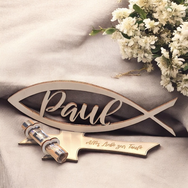 May include: A wooden fish-shaped sign with the name "Paul" engraved on it. The sign is decorated with a small wooden cross and the text "All's Love for Baptism".