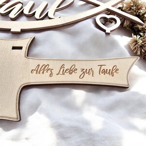 May include: A wooden sign with the text "Alles Liebe zur Taufe" engraved on it. The sign is in the shape of a banner with a pointed end.
