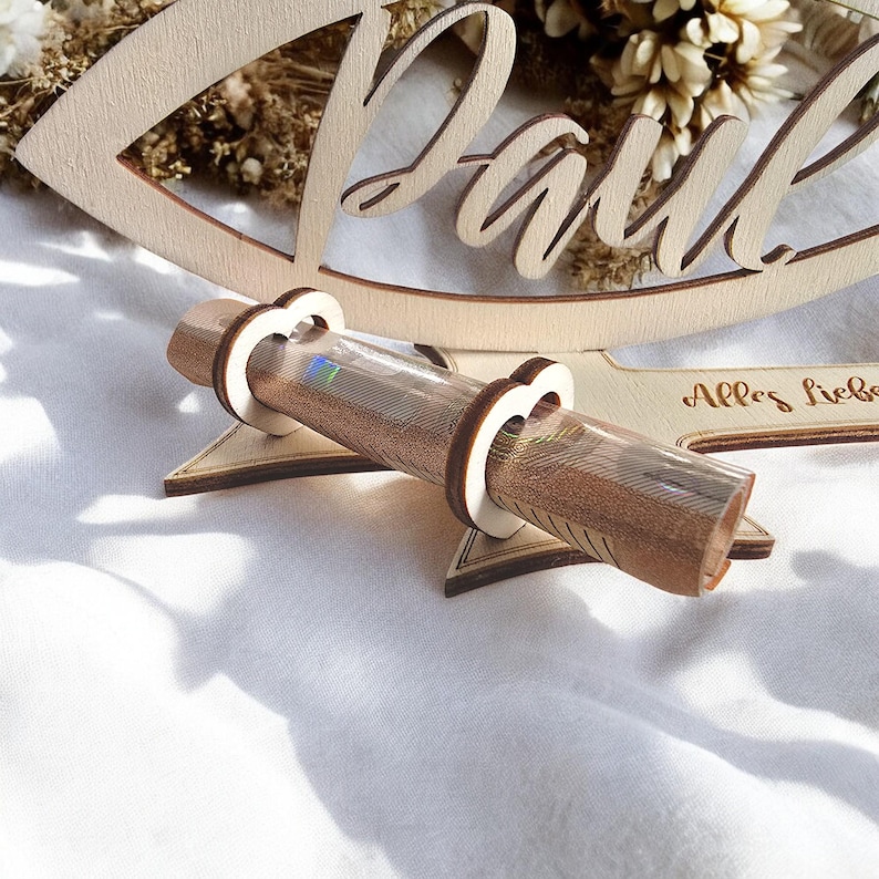 May include: A wooden money holder with the word "Paul" carved into it. The holder is shaped like a fish and has a slot for a rolled bill. The text "Alles Liebe" is also carved into the wood.