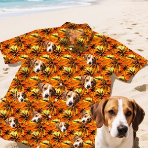 May include: Orange short-sleeved button-down shirt with a repeating pattern of beagle dog faces and palm trees. The shirt has a tropical sunset design. The shirt is on a sandy beach with the ocean in the background.