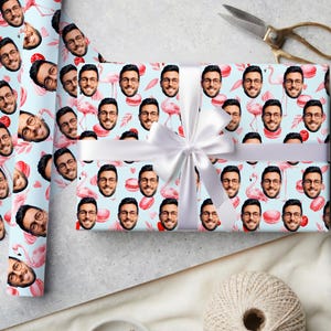Puede incluir: Papel de regalo con un patrón repetido de caras, flamencos y macarons sobre un fondo azul claro. Una cinta y un lazo blancos están atados alrededor del regalo envuelto. Se ven unas tijeras.