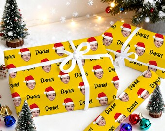 May include: Yellow wrapping paper with a repeating pattern of a child's face wearing a Santa hat and the name "Daniel". The wrapped gifts are tied with white ribbon and surrounded by Christmas decorations, including miniature trees and ornaments.