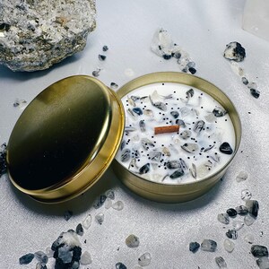 May include: A gold candle jar with a white lid, filled with a white candle and embedded with small black and clear stones. The candle is surrounded by small black and clear stones.