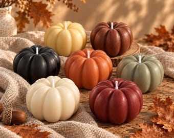 May include: A collection of seven pumpkin-shaped candles in various colors including cream, black, yellow, orange, sage green, brown, and deep red. Each candle features a central white wick and a smooth, ribbed surface resembling a real pumpkin.