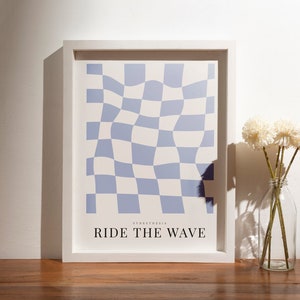 Puede incluir: Una impresión enmarcada con un patrón de cuadros azules y blancos y el texto "RIDE THE WAVE" en negro. La impresión está sobre una superficie de madera con un jarrón de flores blancas en primer plano.