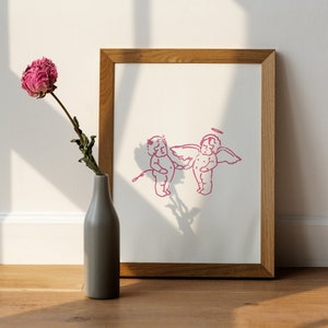 Puede incluir: Una impresión enmarcada de un dibujo lineal rosa de dos querubines con alas. La impresión está en un marco de madera y está sobre un suelo de madera. Una flor rosa seca está en un jarrón gris al lado del marco.
