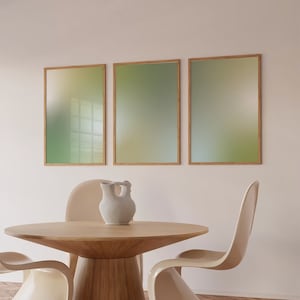 May include: Three framed abstract art prints with a gradient of green and yellow colours hang on a white wall above a wooden table with two white chairs. A white ceramic jug sits on the table.