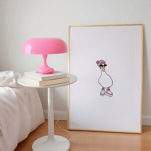 Op de afbeelding: Een wit ingelijste print van een cartoon eend met een zonnebril en een roze muts. De eend draagt roze schoenen en staat op een witte achtergrond. De print hangt aan een witte muur naast een roze lamp op een witte bijzettafel.