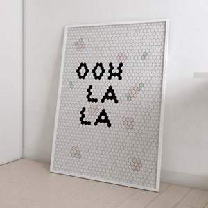 Op de afbeelding: Een witte geframed poster met een wit en zwart honingraatpatroon. De poster zegt "ooh la la" in zwarte blokletters. Er zijn kleine roze, groene en gele bloemen verspreid over het patroon.