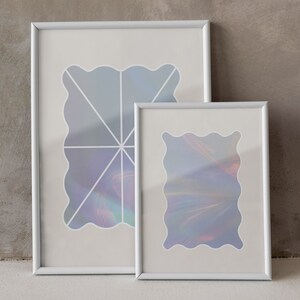 Op de afbeelding: Twee witte fotolijsten met iriserende abstracte ontwerpen erin. De lijsten staan op een grijs, getextureerd oppervlak.