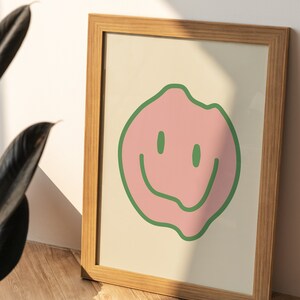 Op de afbeelding: Een ingelijst print met een roze smiley-gezicht omlijnd met groen op een crèmekleurige achtergrond. De lijst is gemaakt van lichtbruin hout.