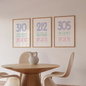 以下が含まれることがあります： 薄い木製のフレームが付いた3つの額装されたプリント。各プリントには薄い青色の大きな数字が描かれており、その下に都市名と州名が書かれています。都市名と州名の下に「Travel is the only thing you buy that makes you richer」というテキストが書かれています。プリントは白い壁に掛けられており、その下に木製のテーブルとベージュの椅子が2つあります。