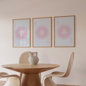 May include: Three framed prints of playing cards with a pink gradient background. The cards are the Queen of Hearts, Queen of Diamonds, and Queen of Clubs. The prints are hanging on a pale pink wall above a wooden table with three chairs.