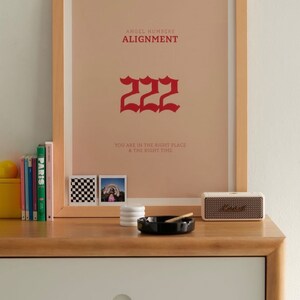 以下が含まれることがあります： ベージュの背景に赤い文字で「ANGEL NUMBERS ALIGNMENT 222 YOU ARE IN THE RIGHT PLACE & THE RIGHT TIME」と書かれた額装されたプリント。プリントは白い壁にかけられており、その下には木製ドレッサーがあり、小さなスピーカー、黒い灰皿、白いコースターが置かれている。