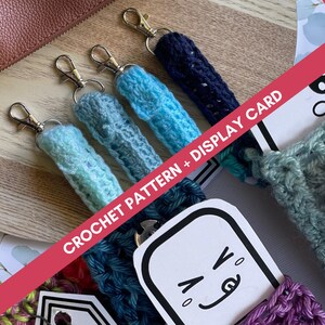 May include: Five crocheted keychains in different colours, including blue, teal, and purple, with silver key rings. The keychains are displayed on a wooden surface with a white card that says "Crochet Pattern + Display Card".