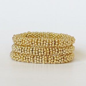 May include: Three gold beaded bracelets stacked on top of each other. The bracelets are made of small, round beads that are tightly packed together.