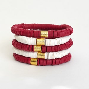 May include: A stack of four bracelets made of red and white beads with gold accents. The bracelets are stacked on top of each other, with the red beads on the outside and the white beads on the inside.