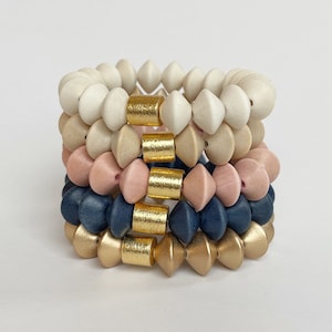 May include: A stack of four wooden bracelets with gold accents. The bracelets are in shades of white, beige, pink, and blue.