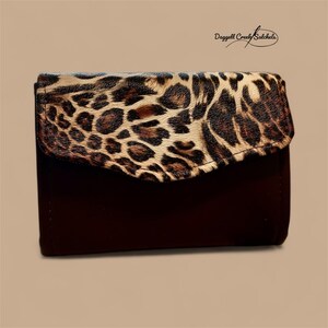 May include: A dark brown wallet with a flap featuring a leopard print design. The wallet has a rectangular shape and a curved flap closure. The text "Daggett Creek Satchels" is in the upper right corner.