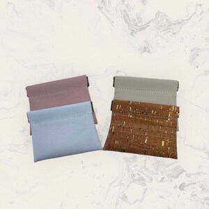 May include: Four small pouches in different colors and materials. The pouches are made of light pink, light blue, light gray, and cork with gold flecks.