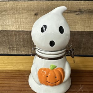 May include: A white ceramic Halloween-themed jar shaped like a ghost, with black eyes and mouth. The ghost holds an orange pumpkin with a green stem. The jar has a metal clasp and a lid.