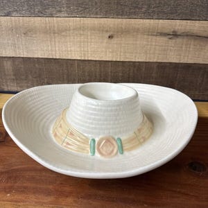 Peut inclure: Plat de service blanc en céramique en forme de sombrero, avec une section centrale surélevée en forme de chapeau. Le bandeau est beige avec des accents roses et verts. Le plat a un design texturé et nervuré.